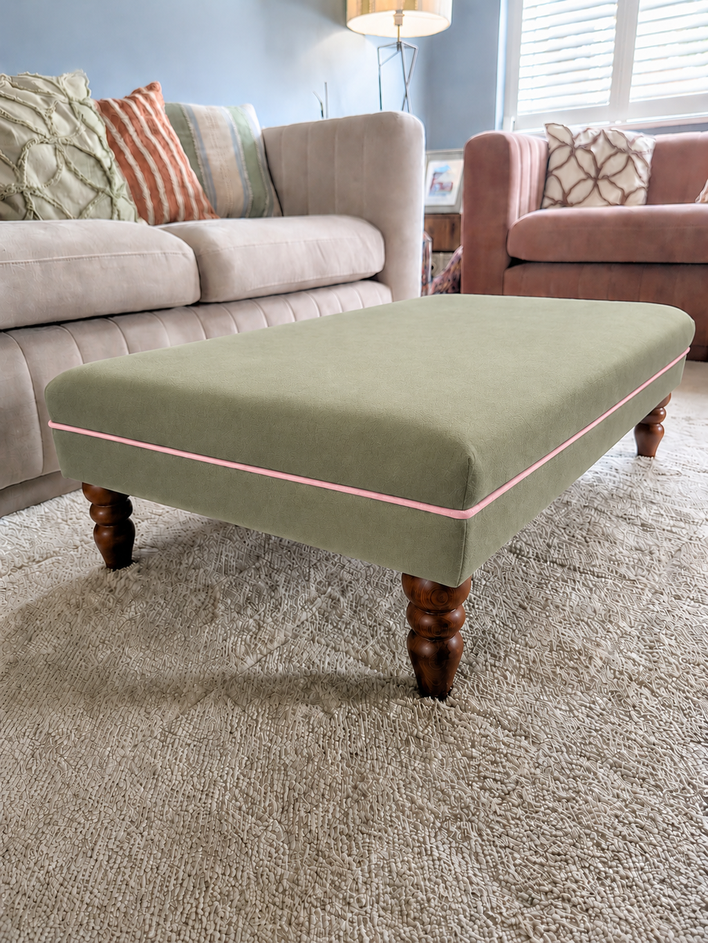 A square upholstered ottoman in sage green with a thin pink piping detail and dark wooden legs, positioned on a textured beige carpet in a living room setting.