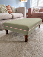 A square upholstered ottoman in sage green with a thin pink piping detail and dark wooden legs, positioned on a textured beige carpet in a living room setting.