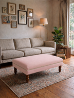 A light pink upholstered rectangular ottoman with turned wooden legs sits on a patterned area rug in a styled living room with a beige sofa, wall art, floor lamp, and plant.