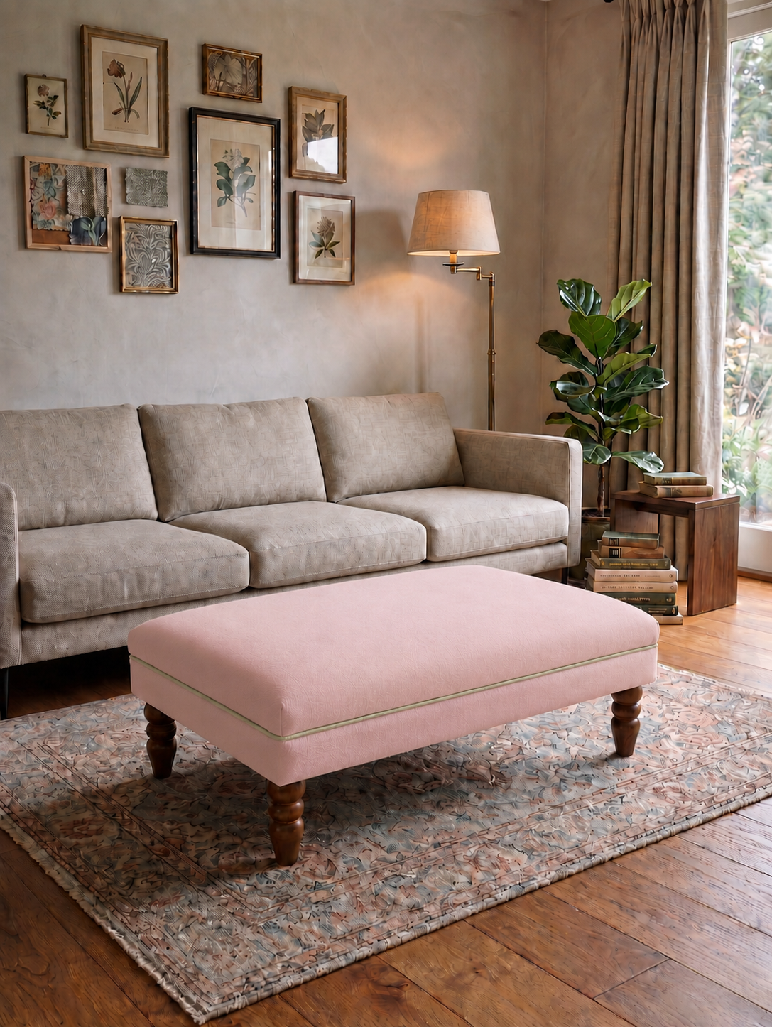 A light pink upholstered rectangular ottoman with turned wooden legs sits on a patterned area rug in a styled living room with a beige sofa, wall art, floor lamp, and plant.