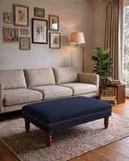 A styled living room featuring a beige fabric sofa, a dark navy blue rectangular ottoman with wooden legs and piping, a patterned area rug, a floor lamp, and a potted plant.