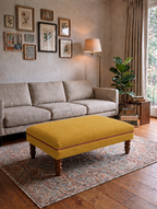A mustard yellow upholstered rectangular ottoman with turned wooden legs sits on a patterned area rug in a styled living room with a beige sofa, wall art, a floor lamp, and a potted plant.
