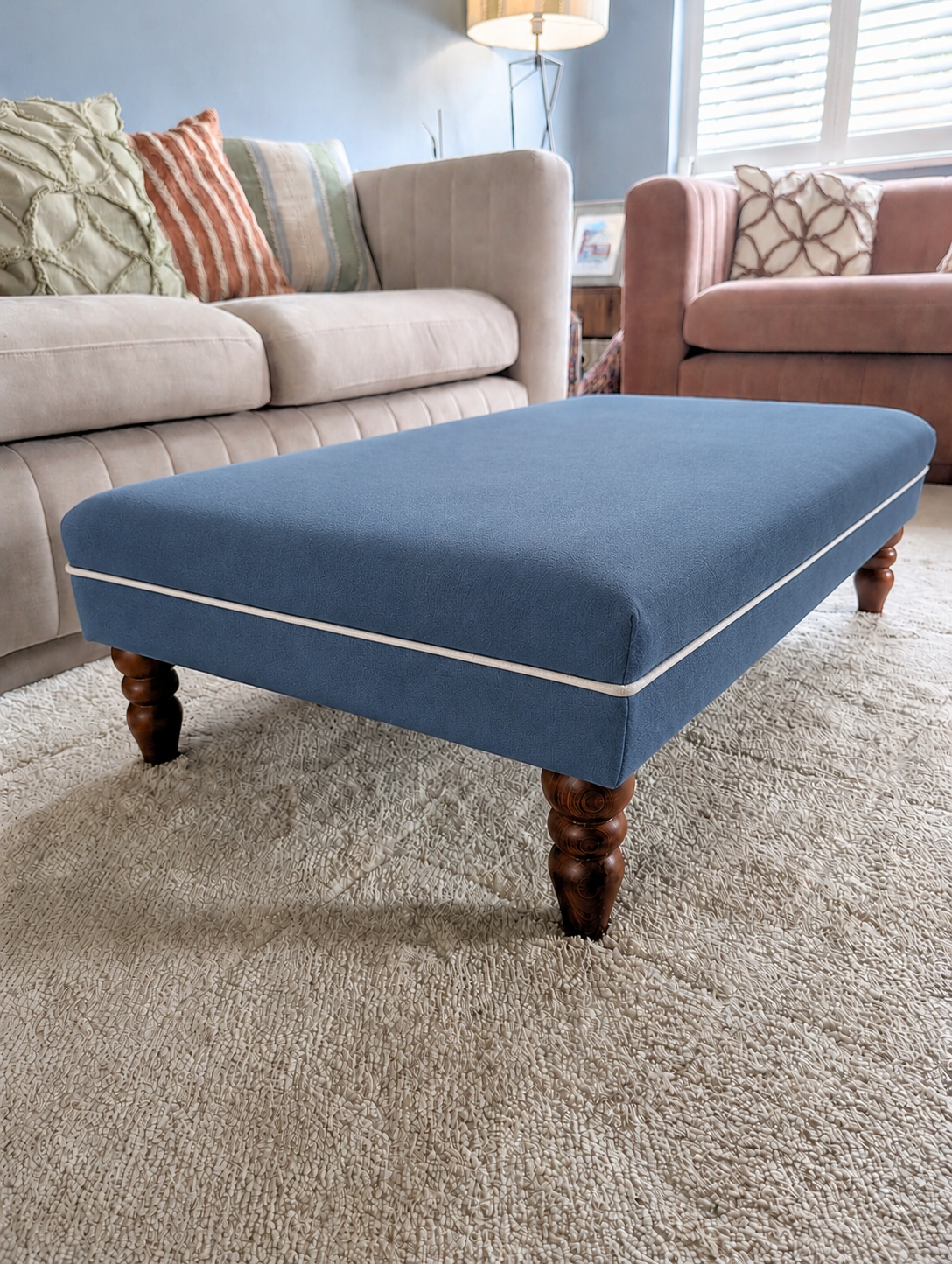 A blue upholstered ottoman with dark wooden legs sits on a beige carpet in a living room, positioned between a cream sofa and a pink armchair.