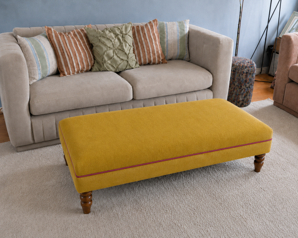 A mustard yellow upholstered coffee table with turned wooden legs and a thin red piping accent, positioned in front of a light beige sofa with decorative throw pillows in a living room setting.