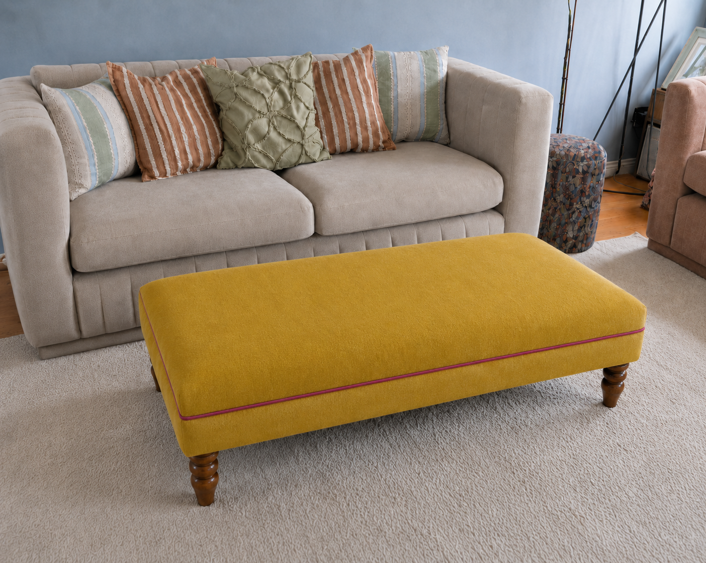 A mustard yellow upholstered coffee table with turned wooden legs and a thin red piping accent, positioned in front of a light beige sofa with decorative throw pillows in a living room setting.