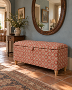 An upholstered storage bench with a terracotta and cream floral pattern, wooden legs, and a hinged lid, set in a styled entryway with a blue wall, large mirror, and terracotta tile floor.
