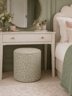 A round upholstered ottoman stool with a green and pink floral pattern by Ian Sanderson, sits under a white vanity table in a styled bedroom setting.