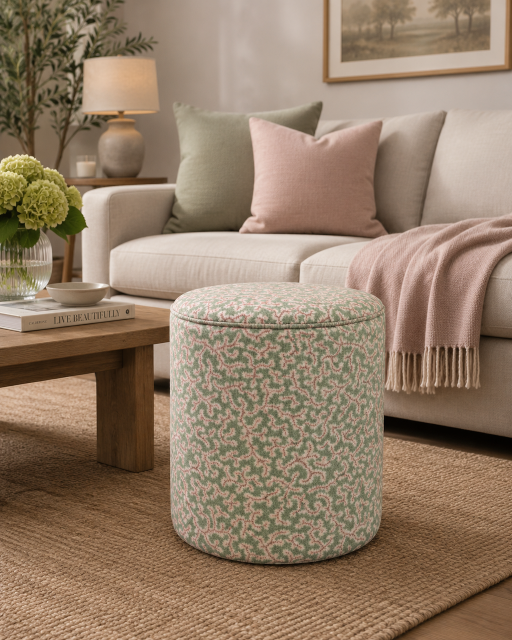A cylindrical upholstered ottoman with a green and pink floral pattern sits on a woven rug in a styled living room next to a sofa with neutral-toned pillows and a pink throw blanket.