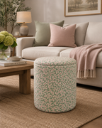 A cylindrical upholstered ottoman with a green and pink floral pattern sits on a woven rug in a styled living room next to a sofa with neutral-toned pillows and a pink throw blanket.