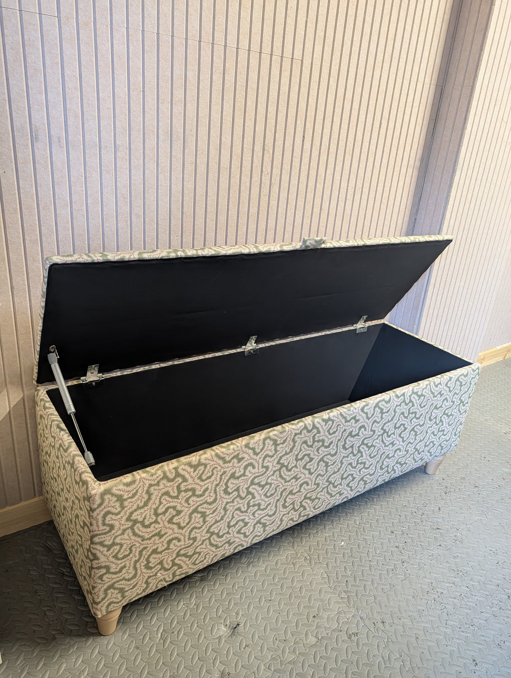 A rectangular upholstered storage bench with its lid open, showing a black fabric-lined interior. The bench has a beige and gray wavy patterned fabric exterior and small wooden legs, set against a light-colored paneled wall and textured gray floor.