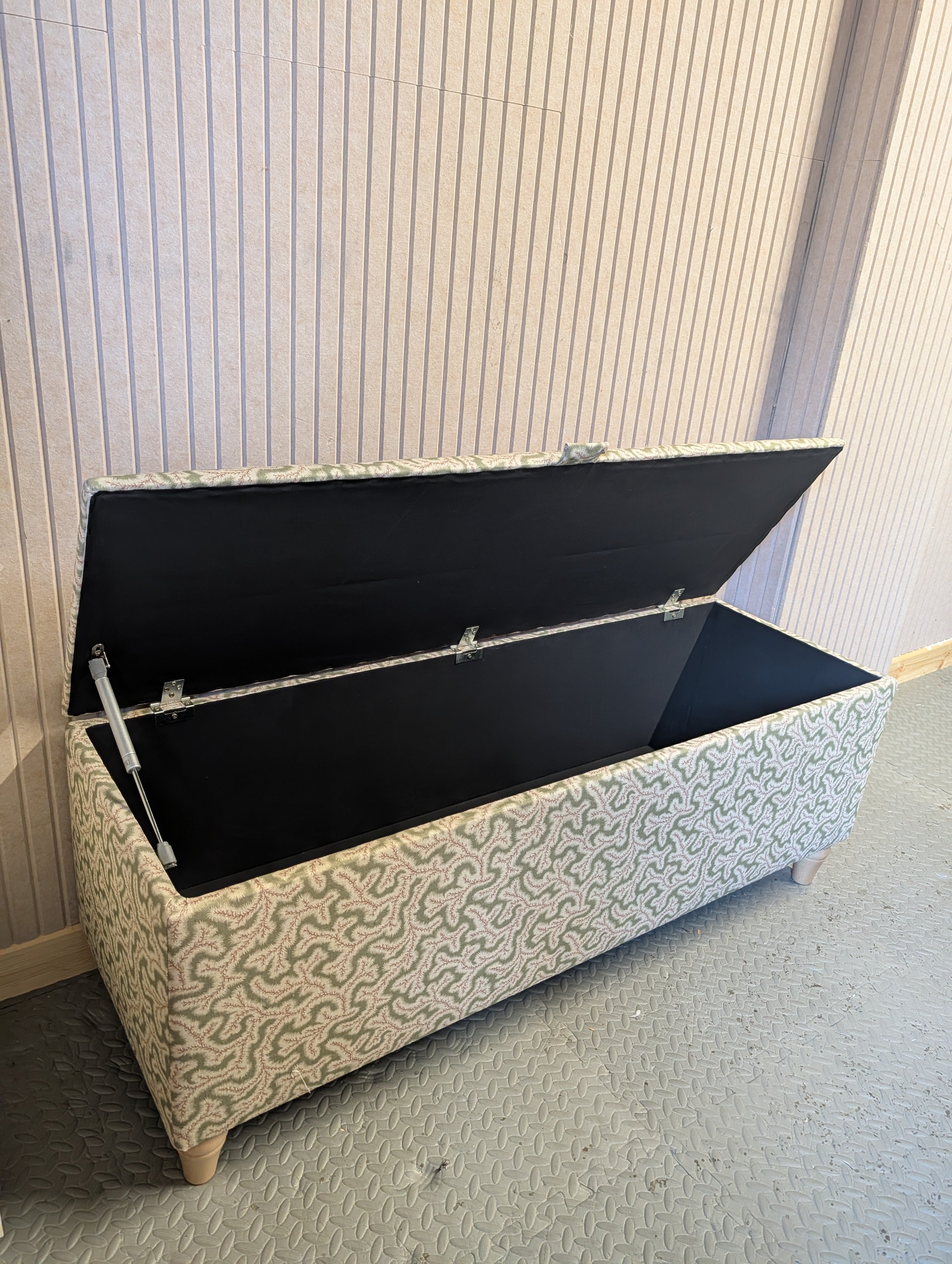 A rectangular upholstered storage bench with its lid open, showing a black fabric-lined interior. The bench has a beige and gray wavy patterned fabric exterior and small wooden legs, set against a light-colored paneled wall and textured gray floor.
