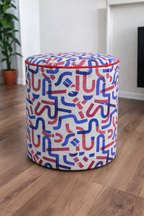 A cylindrical upholstered ottoman stool with a white fabric cover featuring a red and blue abstract maze-like pattern, set on a wooden floor in a room with a potted plant in the background.