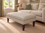 An upholstered rectangular ottoman with a green and beige floral pattern and dark wooden legs, shown in a styled living room next to a cream sofa.