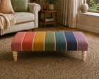 A rectangular upholstered ottoman with turned wooden legs, featuring vertical rainbow-colored stripes in pink, red, orange, yellow, green, teal, blue, and purple separated by thin white lines. It is placed on a textured beige rug in a living room setting with a sofa and side table in the background.