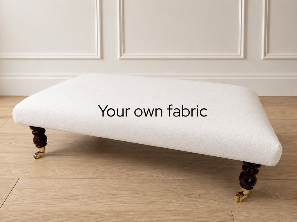 A low upholstered bench or ottoman with a white fabric seat and dark turned wooden legs with gold casters, shown in a room with paneled walls and wood flooring. The words 'Your own fabric' are printed on the seat.