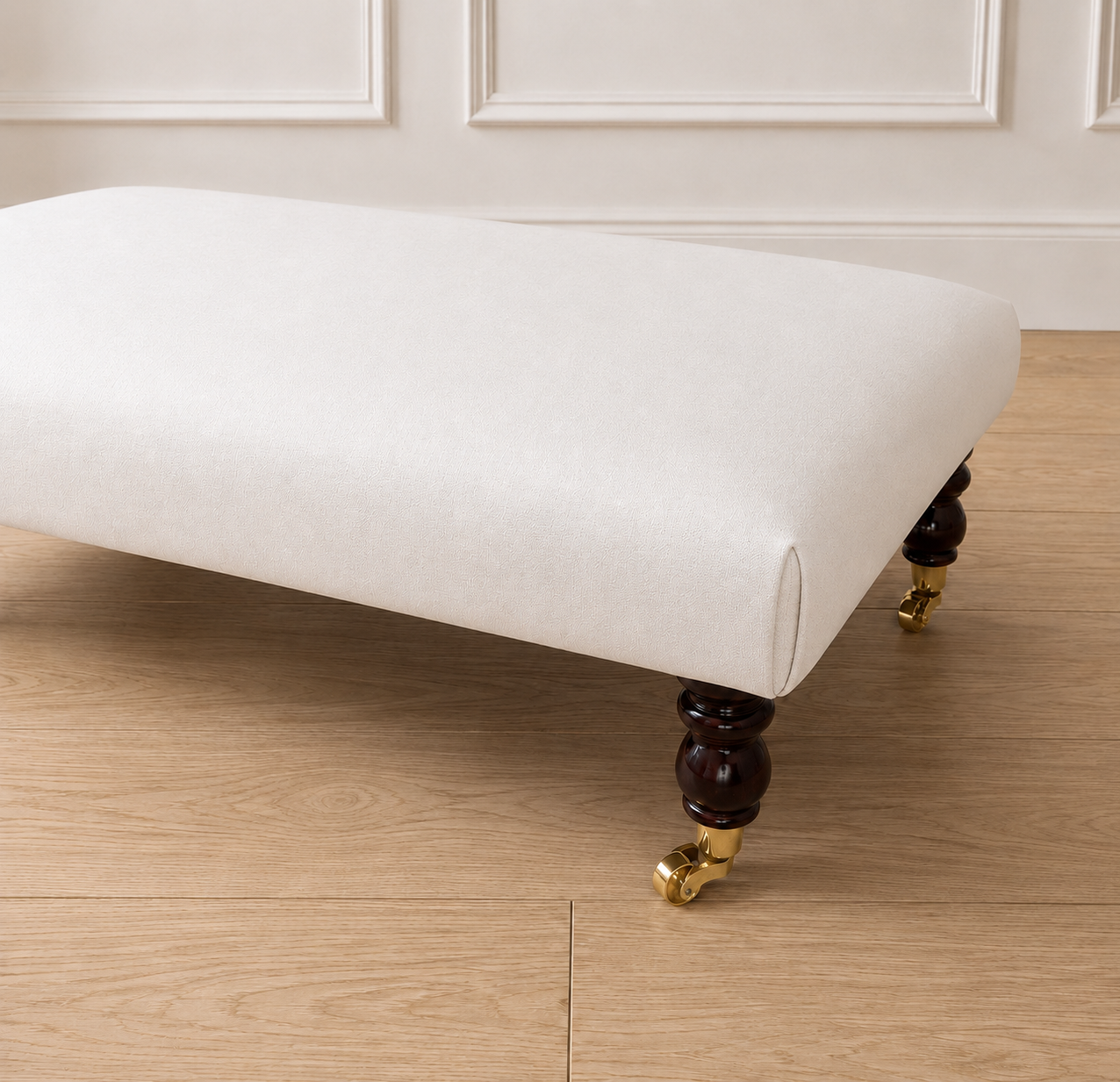 An upholstered bench with a cream fabric seat cushion and turned dark wood legs with gold caster wheels, shown on a light wood floor against a white paneled wall.
