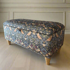 Decorative ottoman with floral pattern on a wooden floor next to a wall with a framed picture.