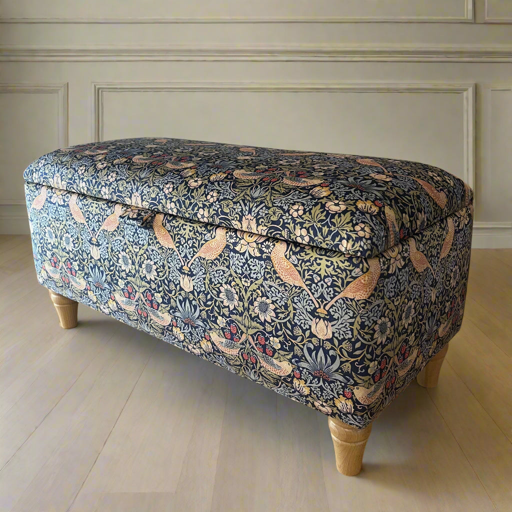 Decorative ottoman with floral pattern on a wooden floor next to a wall with a framed picture.