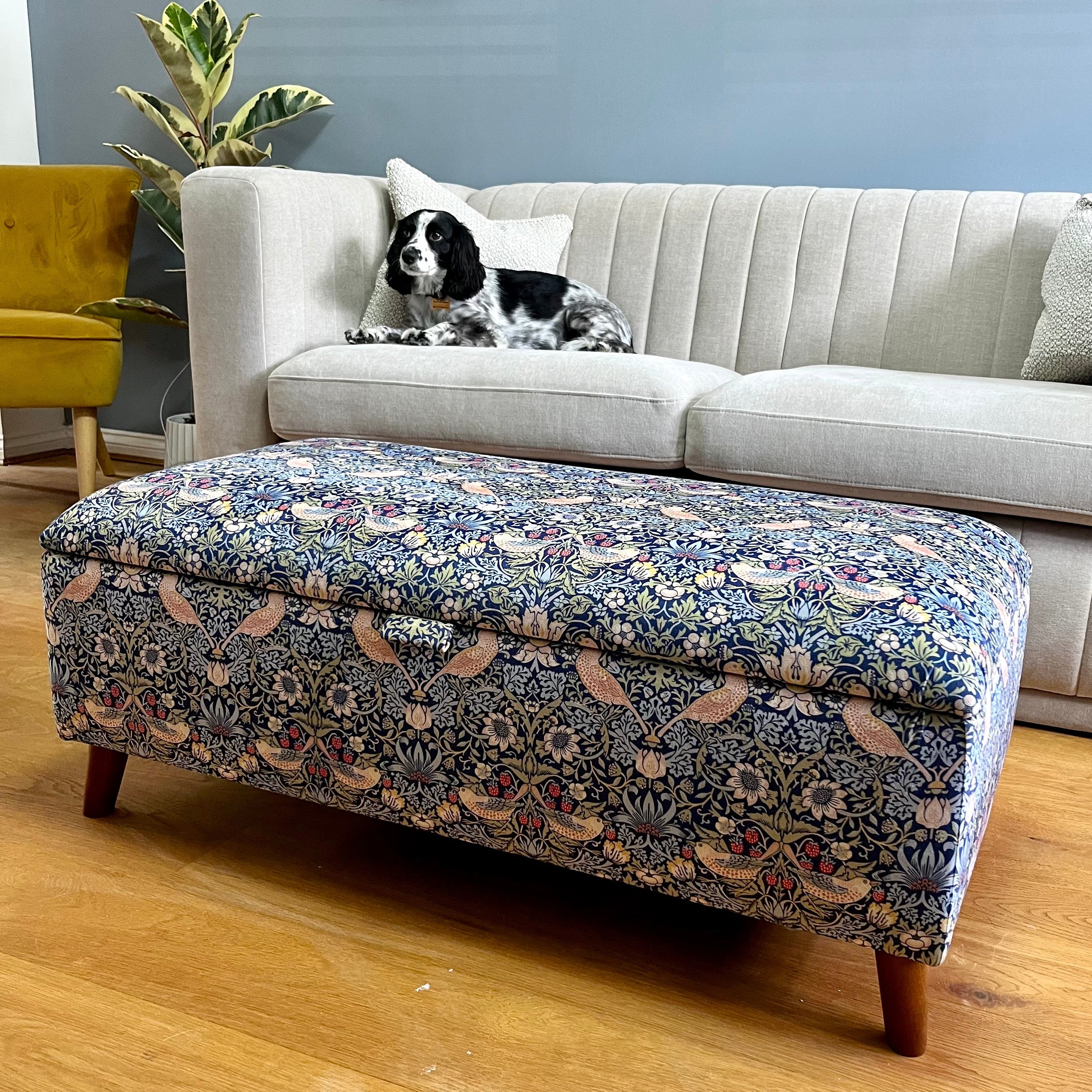 Dog lying on a gray sofa with a floral  Navy storage ottoman footstool in William morris strawberry thief in front