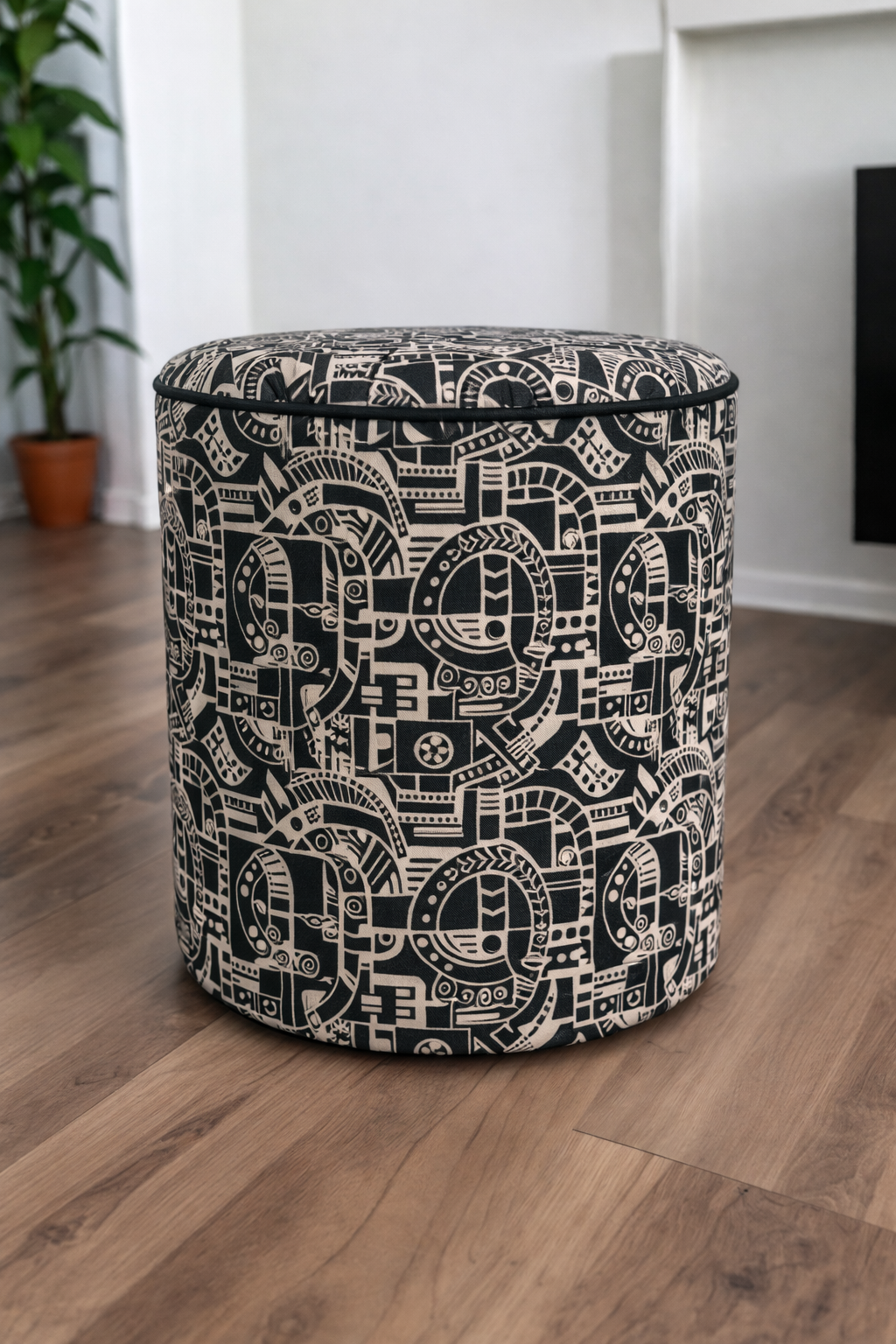 A cylindrical upholstered ottoman or stool with a black and off-white geometric Labyrinth pattern, set on a wooden floor in a modern room.