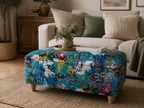 A floral ottoman with a vibrant blue background and multicolored botanical print, styled in a cozy living room setting with a sofa and decorative pillows behind it.