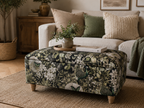 A floral upholstered ottoman with wooden legs sits in a living room setting, styled with a small plant and books on top, with a sofa and decorative pillows in the background.