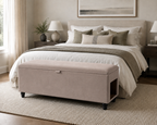 A beige upholstered storage bench with black legs sits and a side pet entrance at the foot of a neatly made bed in a softly lit bedroom. The bed is dressed in white and taupe bedding with matching pillows, and the room includes nightstands, lamps, a rug, and framed wall art.