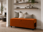 An orange upholstered storage bench with black legs sits and a side pet entrance at the foot of a neatly made bed in a softly lit hallway. The hallway is neutral  in white and beige decor with wooden selves.