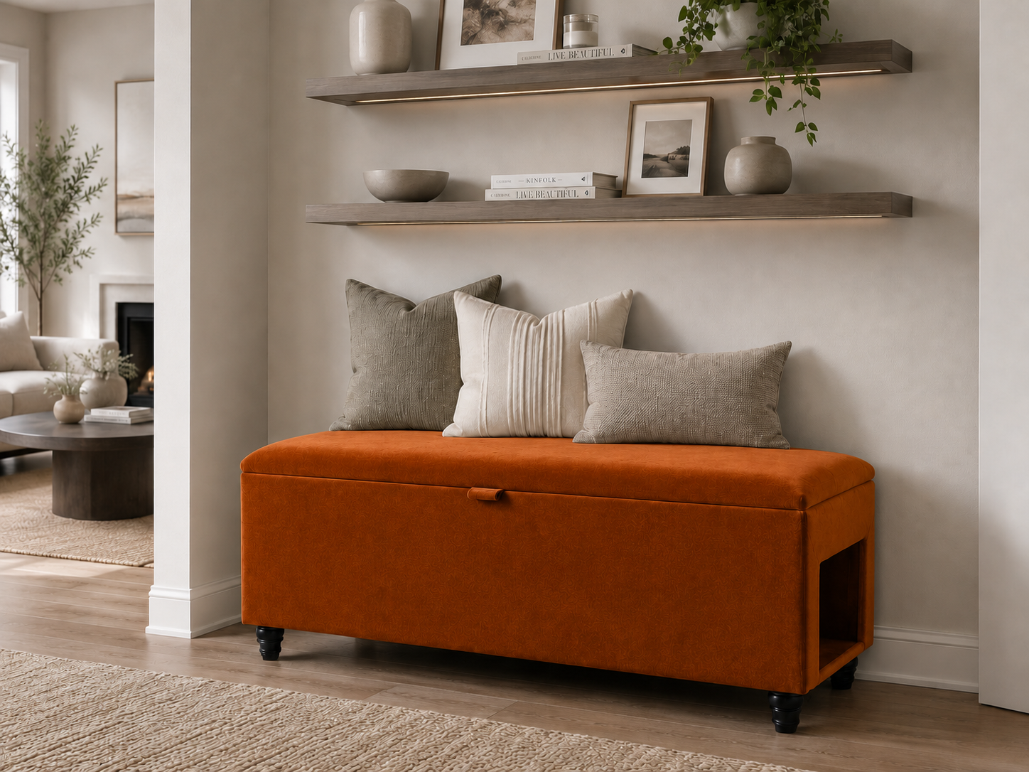 An orange upholstered storage bench with black legs sits and a side pet entrance at the foot of a neatly made bed in a softly lit hallway. The hallway is neutral  in white and beige decor with wooden selves.