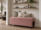 A rose pink upholstered storage bench with black legs sits and a side pet entrance at the foot of a neatly made bed in a softly lit hallway. The hallway is neutral  in white and beige decor with wooden selves.
