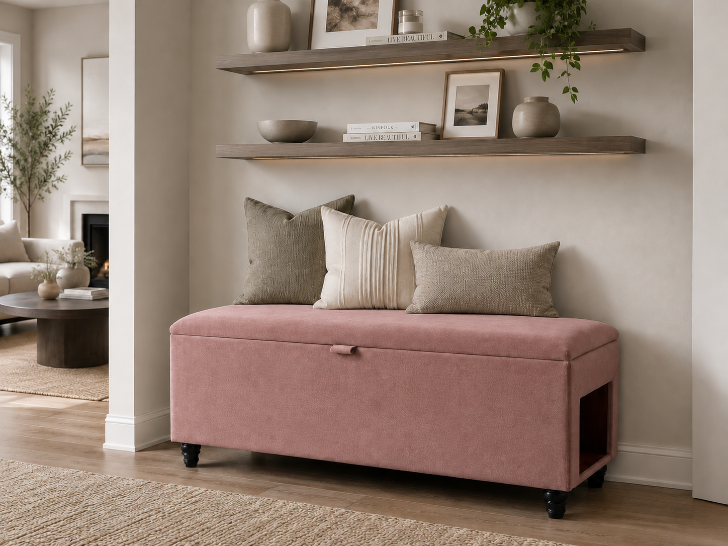 A rose pink upholstered storage bench with black legs sits and a side pet entrance at the foot of a neatly made bed in a softly lit hallway. The hallway is neutral  in white and beige decor with wooden selves.