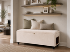 An upholstered storage bench with a cream fabric cover and black casters, styled with three neutral throw pillows in a modern living room setting with floating shelves.