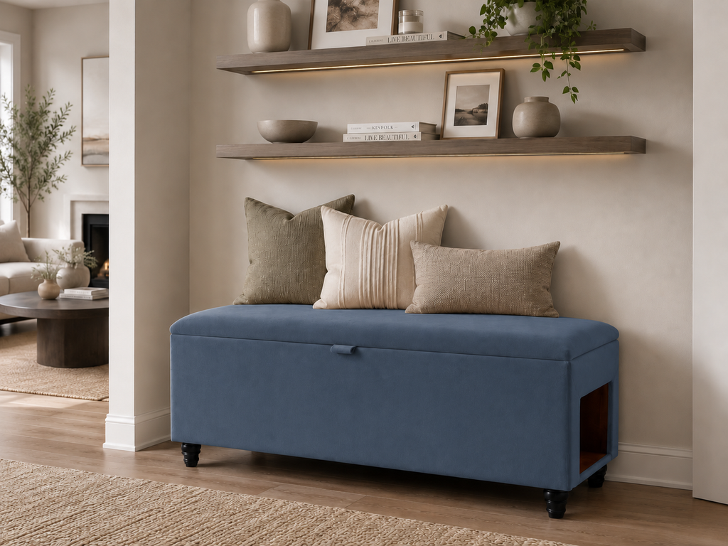 A denim blue upholstered storage bench with black caster wheels, styled with four neutral throw pillows in a modern living room setting with floating shelves above.