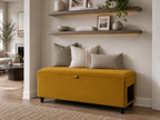 A mustard yellow upholstered storage bench with a small front pull handle and black caster wheels, styled in a living room setting with three neutral throw pillows. Above the bench are two floating wood shelves with decor items.