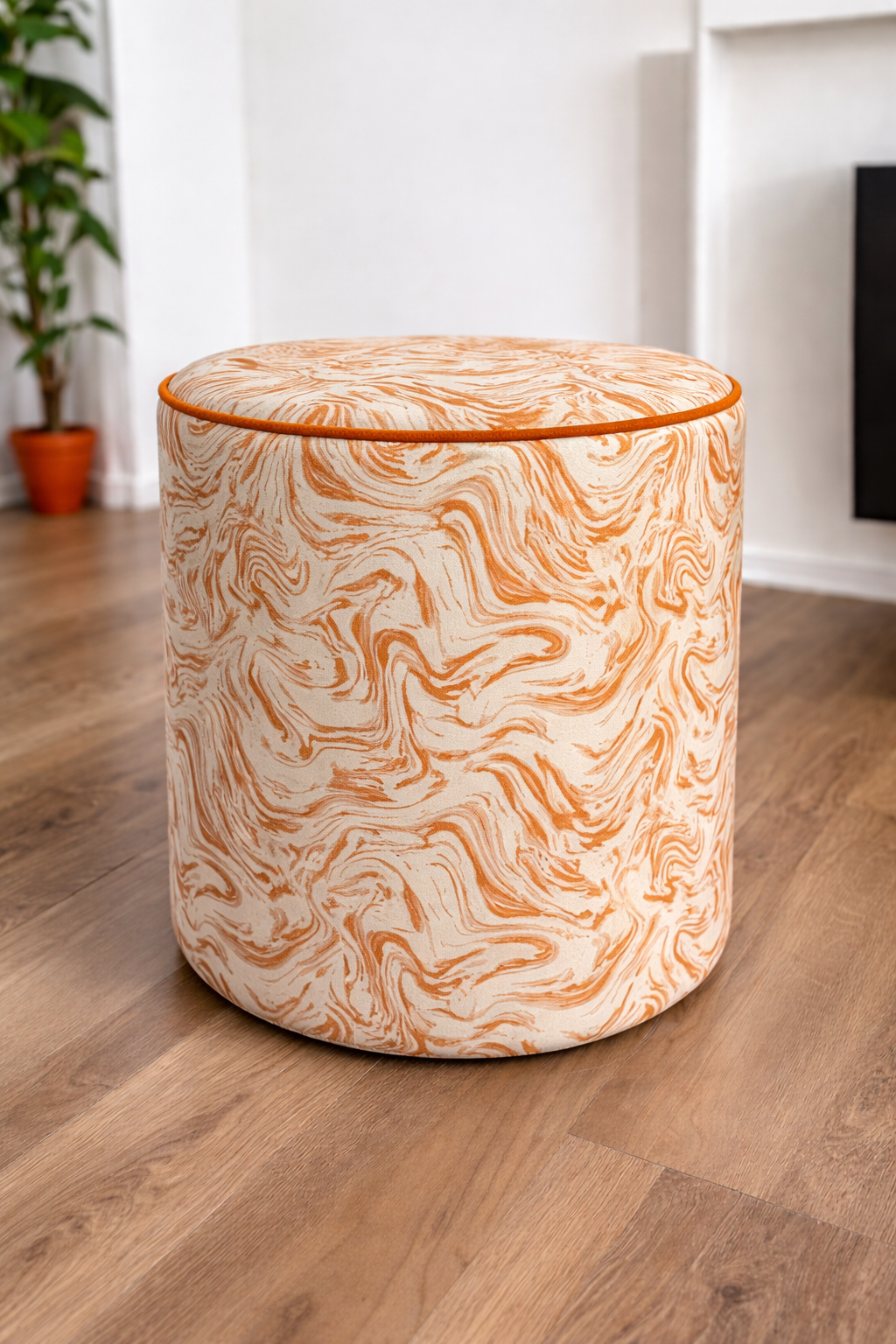 A cylindrical upholstered ottoman or stool with a swirling orange and cream pattern, standing on a light brown wood floor in a bright room.