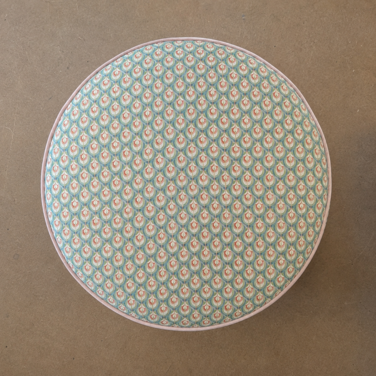 A round upholstered stool or ottoman viewed from above, featuring a light blue and white repeating floral pattern with a thin pinkish-beige outer rim, set against a plain brown background.