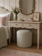 A styled bedroom scene featuring a cream-colored vanity desk with two drawers, a gold-framed mirror, and a matching patterned cylindrical ottoman in a pink and green scallop design. The room has light wood floors, paneled walls, and a bed with white linens and a pink pillow on the left.