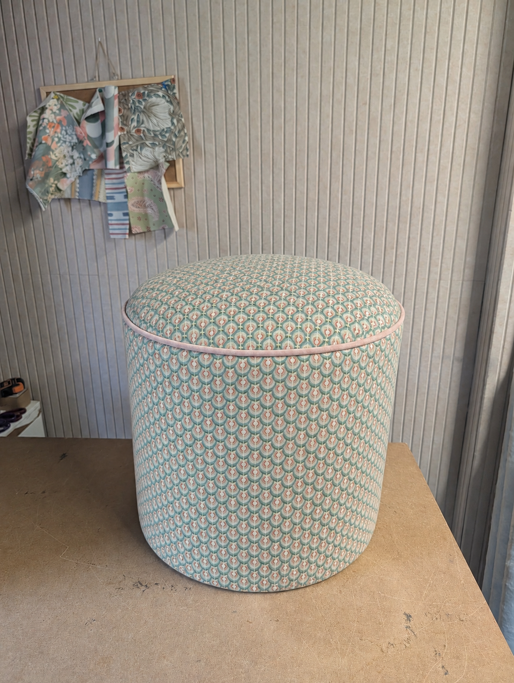 A cylindrical upholstered ottoman or stool with a light green, pink, and white scalloped pattern, sitting on a wooden table against a paneled wall.