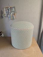 A cylindrical upholstered ottoman or stool with a light green, pink, and white scalloped pattern, sitting on a wooden table against a paneled wall.
