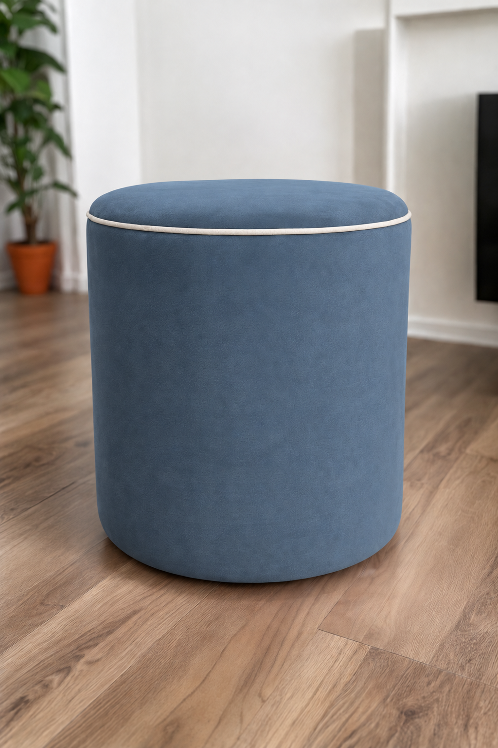 A round upholstered ottoman stool in a denim velvet with cream piping fabric, set on a light brown wood floor in a modern room with white walls and a potted plant in the background.