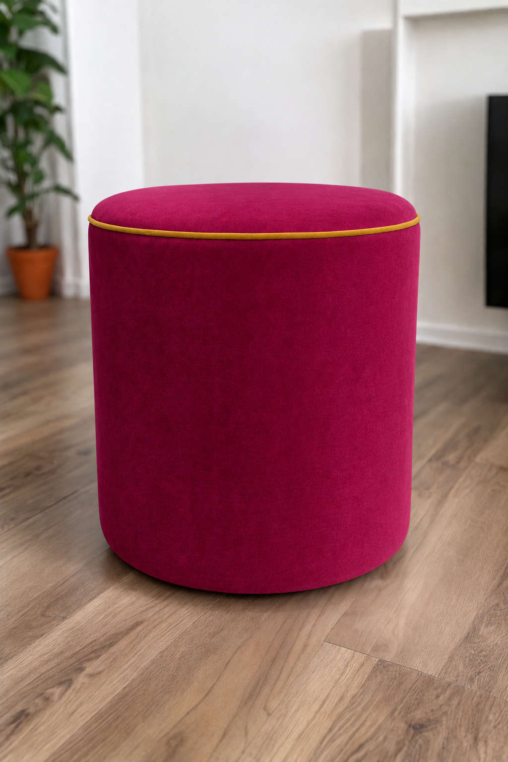 A round upholstered ottoman stool in a raspberry pink velvet fabric with contrasting honey piping, set on a light brown wood floor in a modern room with white walls and a potted plant in the background.