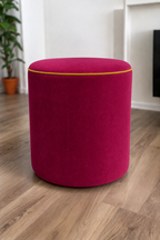 A round upholstered ottoman stool in a raspberry pink velvet fabric with contrasting honey piping, set on a light brown wood floor in a modern room with white walls and a potted plant in the background.
