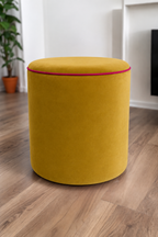 A round upholstered ottoman stool in a honey yellow velvet fabric with contrasting raspberry piping, set on a light brown wood floor in a modern room with white walls and a potted plant in the background.