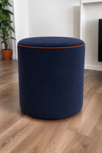A round upholstered ottoman stool in a royal blue velvet fabric with amber piping, set on a light brown wood floor in a modern room with white walls and a potted plant in the background.