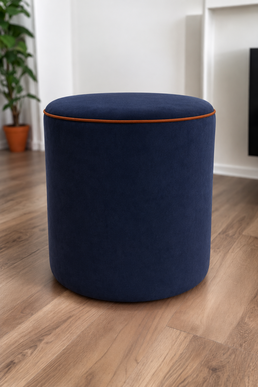 A round upholstered ottoman stool in a royal blue velvet fabric with amber piping, set on a light brown wood floor in a modern room with white walls and a potted plant in the background.