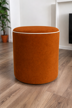 A round upholstered ottoman stool in an amber velvet fabric with cream piping, set on a light brown wood floor in a modern room with white walls and a potted plant in the background.