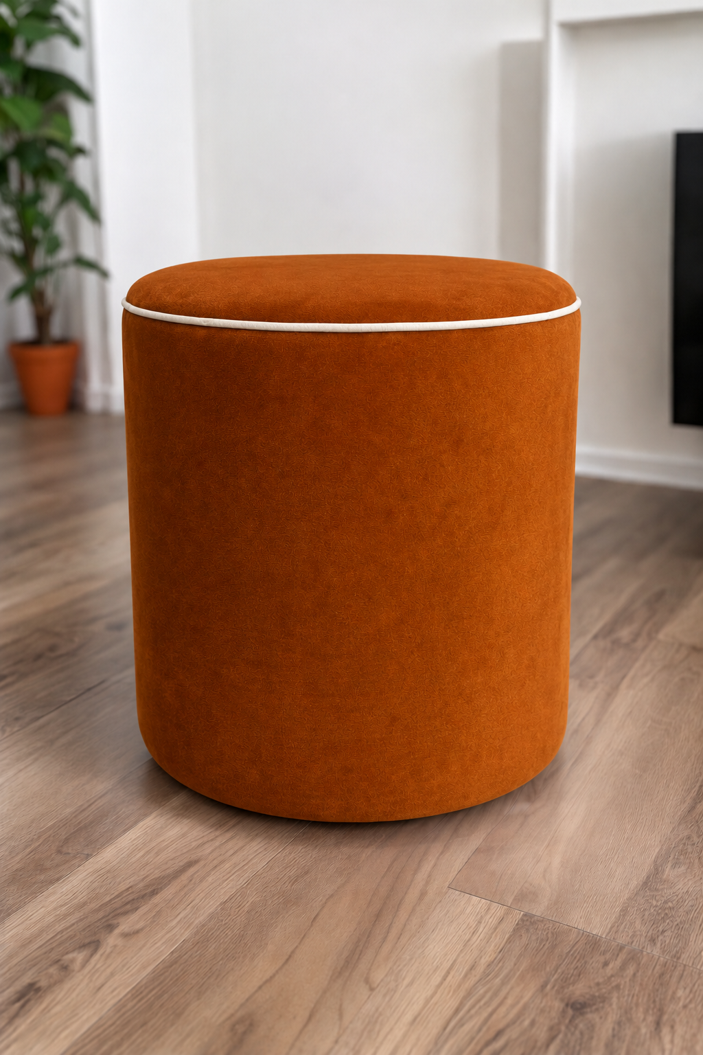 A round upholstered ottoman stool in an amber velvet fabric with cream piping, set on a light brown wood floor in a modern room with white walls and a potted plant in the background.
