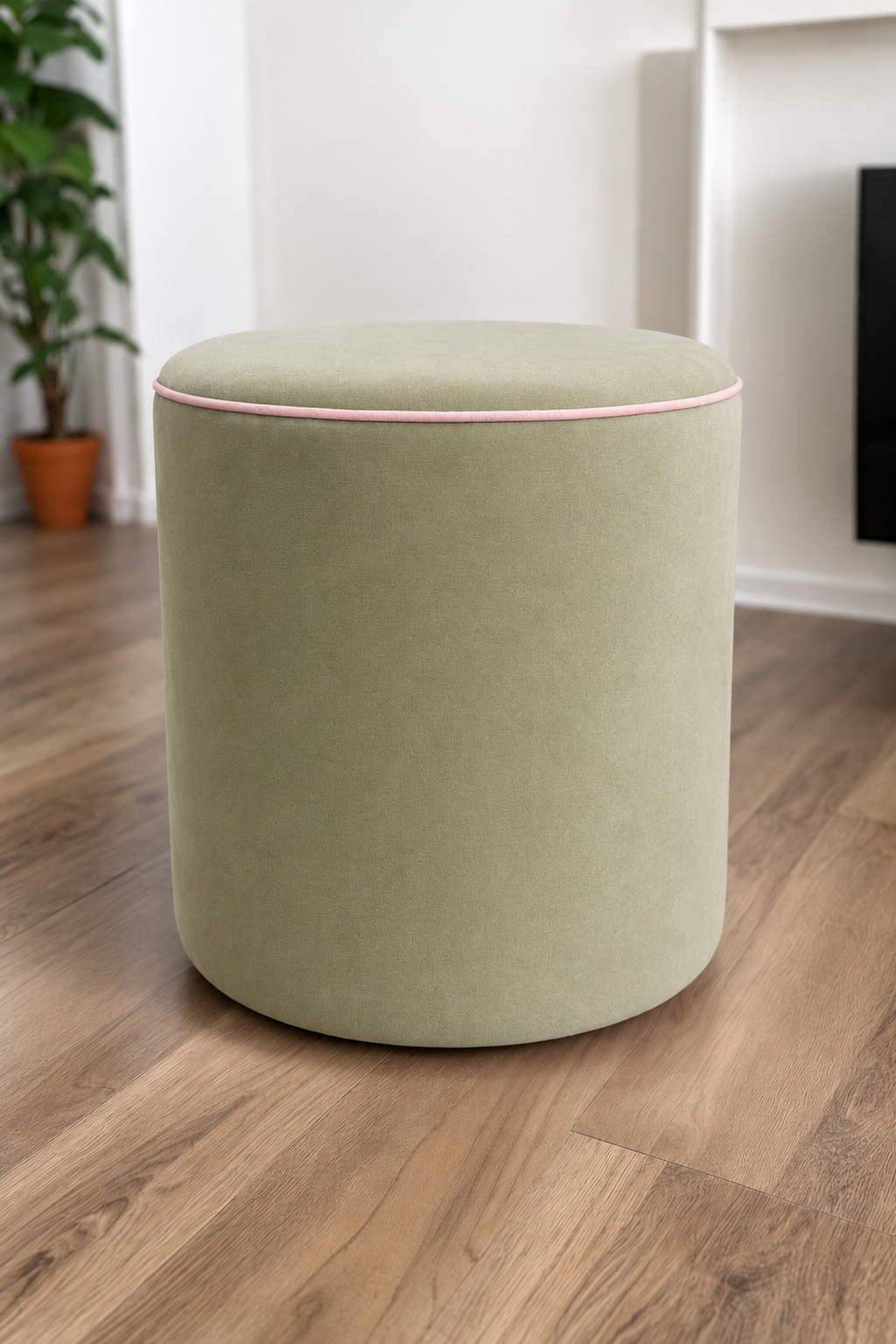 A cylindrical upholstered ottoman in sage green velvet with a thin pink piping trim along the top edge, set on a light brown wooden floor in a modern room.