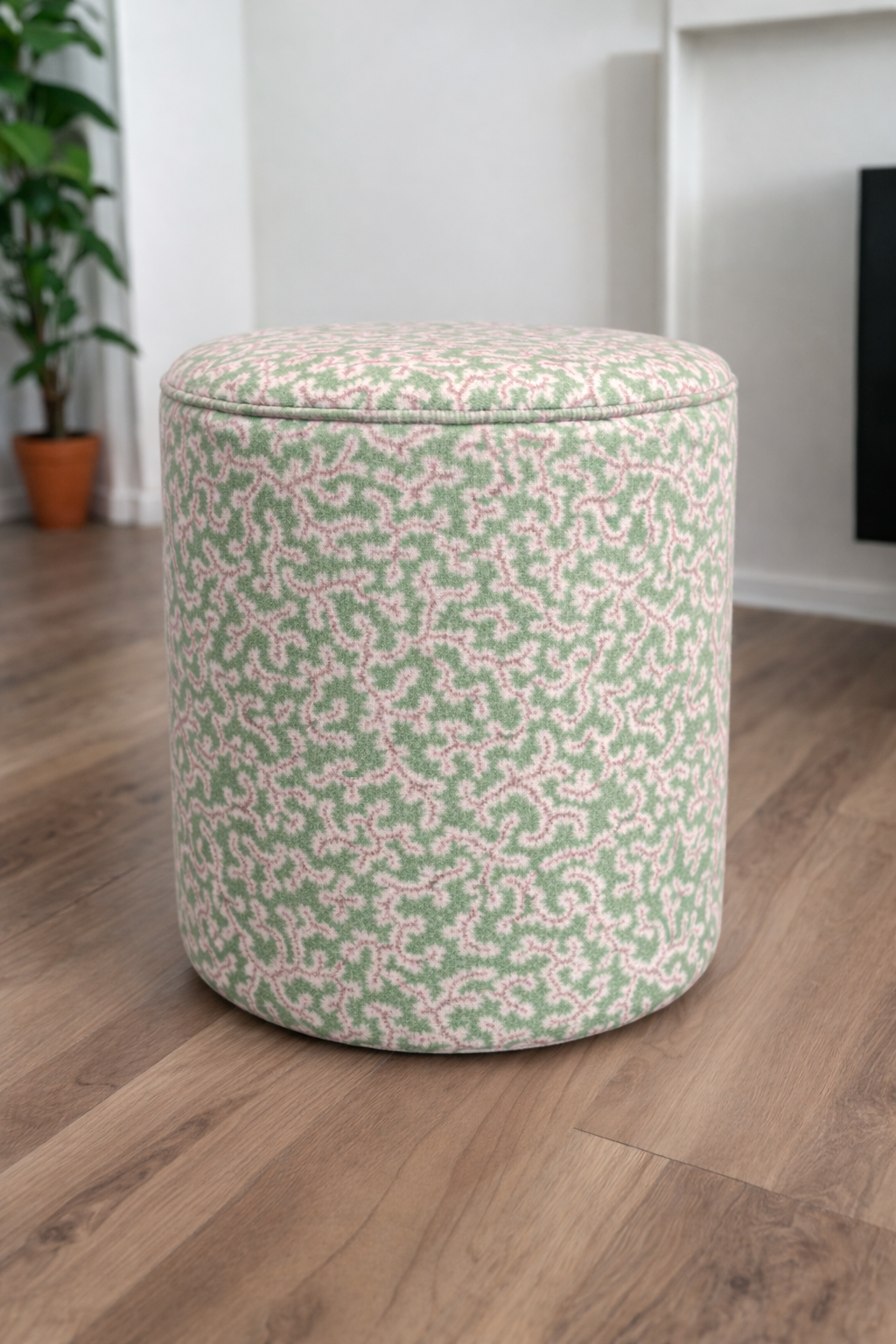 A round upholstered ottoman stool with a light green and pink floral pattern, set on a wooden floor in a bright room.