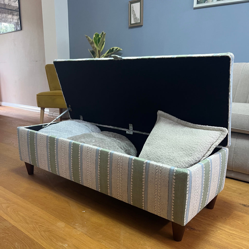Green, blue and white striped ottoman with walnut legs, open with blankets and cushions inside.
